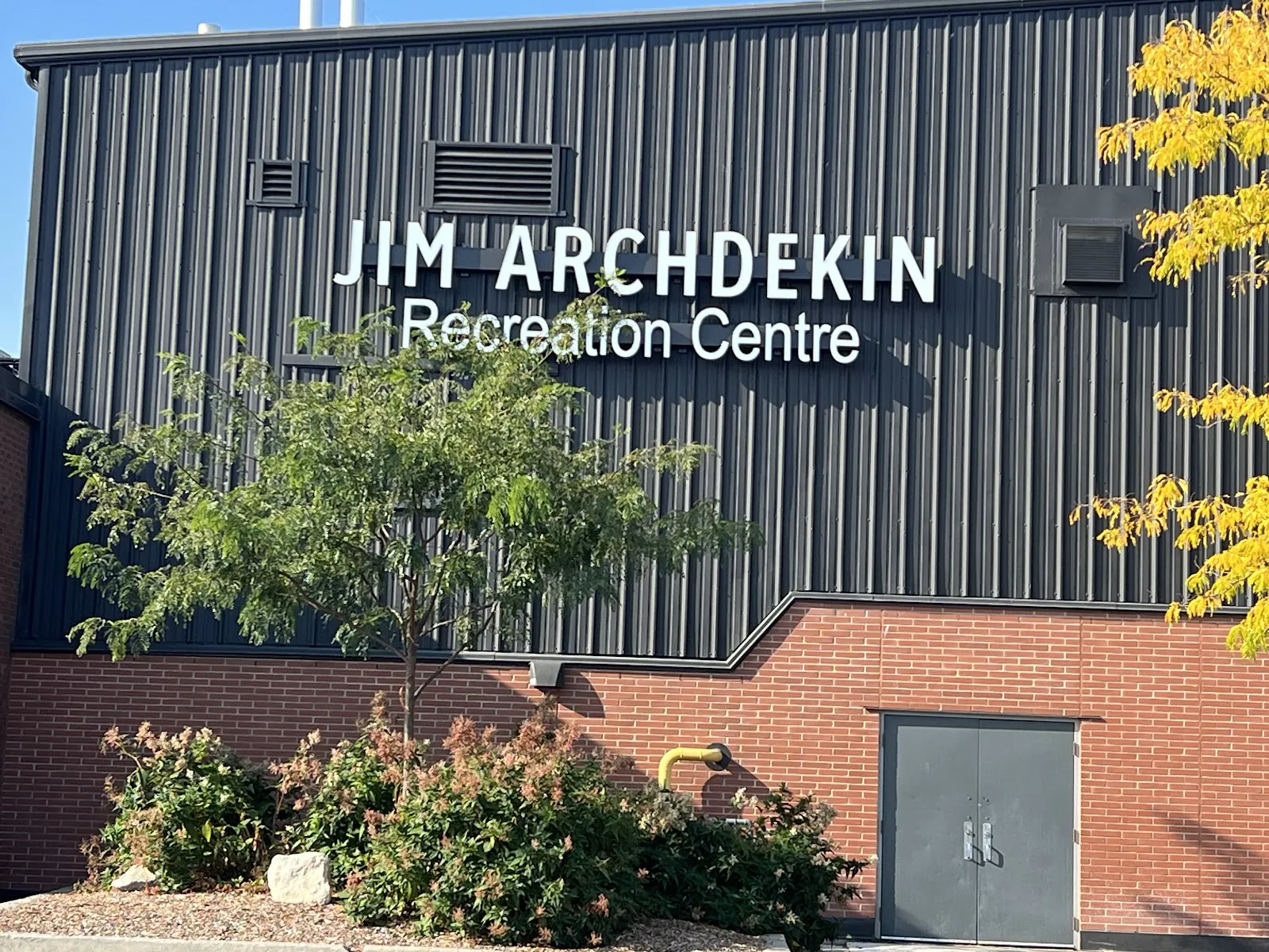 Muro del edificio con revestimiento metálico oscuro y las letras "Jim Archdekin Recreation Centre" sobre una base de ladrillo, con una puerta de servicio y arbustos al frente. — Jim Archdekin Recreation Centre