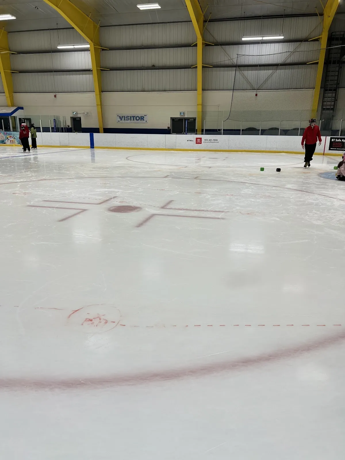 Círculos de saque y marcas sobre el hielo en el Jim Archdekin Recreation Centre, con arcos de acero amarillos, el letrero para visitantes y algunos patinadores a lo largo de las vallas del fondo. — Jim Archdekin Recreation Centre