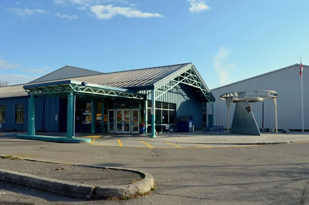 The recreation centre's blue-trimmed metal entrance canopy and glass doors, with the larger arena building and a metal sculpture visible beside the parking lot. — Jim Durrell Recreation Centre