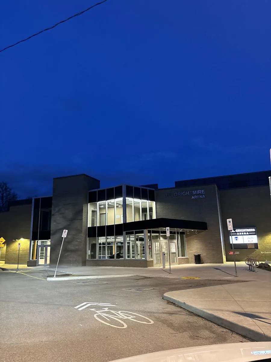 La entrada renovada con fachada de vidrio de la arena al anochecer, iluminada desde dentro, con la marca de un carril para bicicletas en primer plano. — J.L. Grightmire Arena