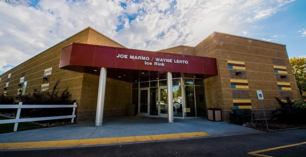 The arena's tan brick entrance with a red canopy reading "Joe Marmo / Wayne Lehto Ice Rink" over glass doors, white split-rail fencing and trees to the left. — Joe Marmo / Wayne Lehto Ice Arena