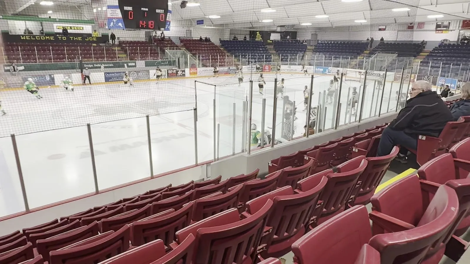 A wide view along rows of red seats, the sheet and a fan-filled far side under a steel roof with a long window wall above. — Joe Thornton Community Centre