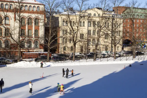 The Johanneksenkenttä artificial ice field in Helsinki in winter, with skaters and children on the ice and apartment buildings behind. — Johanneksenkenttä – tekojääkenttä