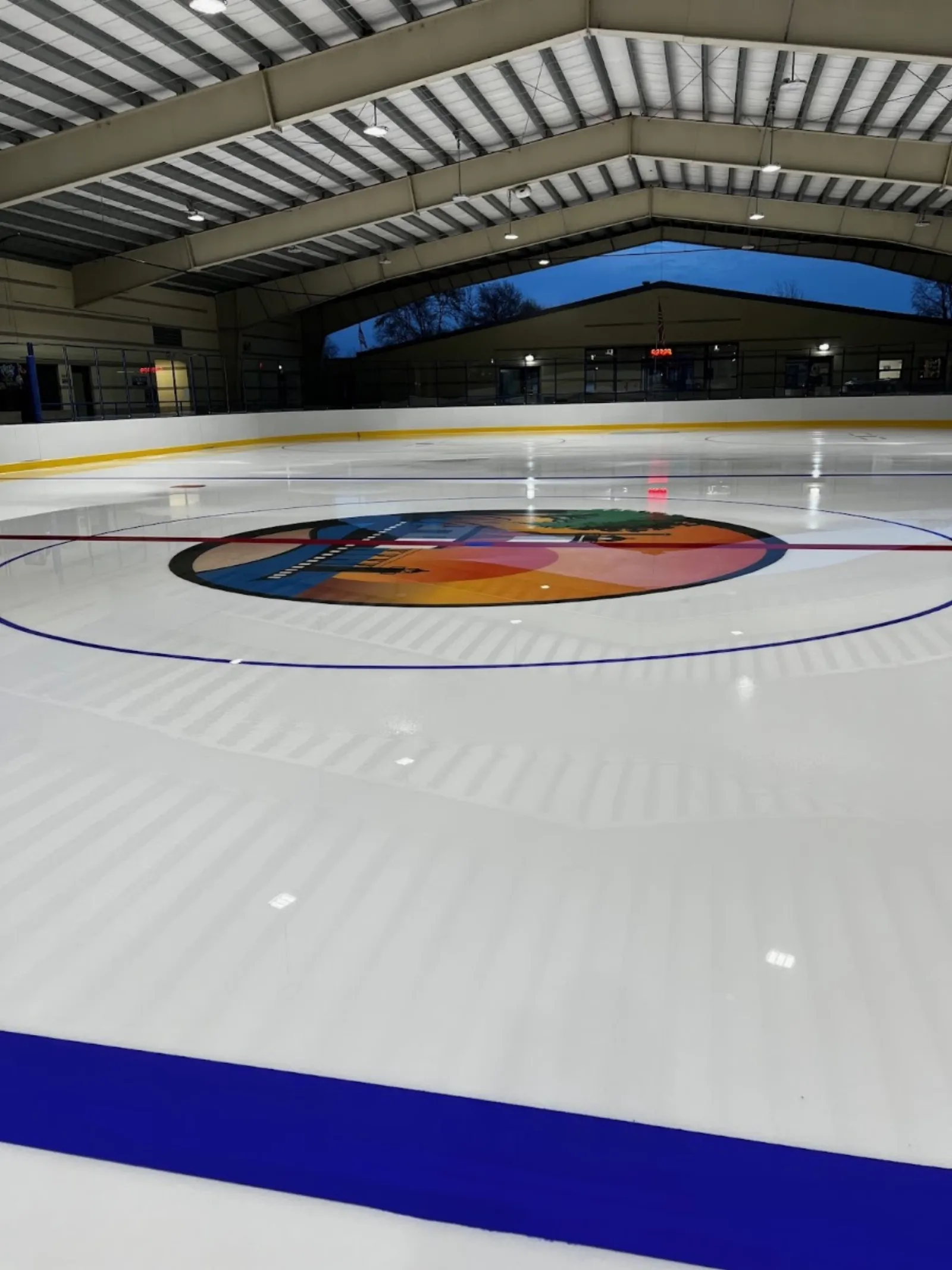 The empty ice with its center-ice logo under the open-sided steel roof. — John & Dede Howard Ice Arena