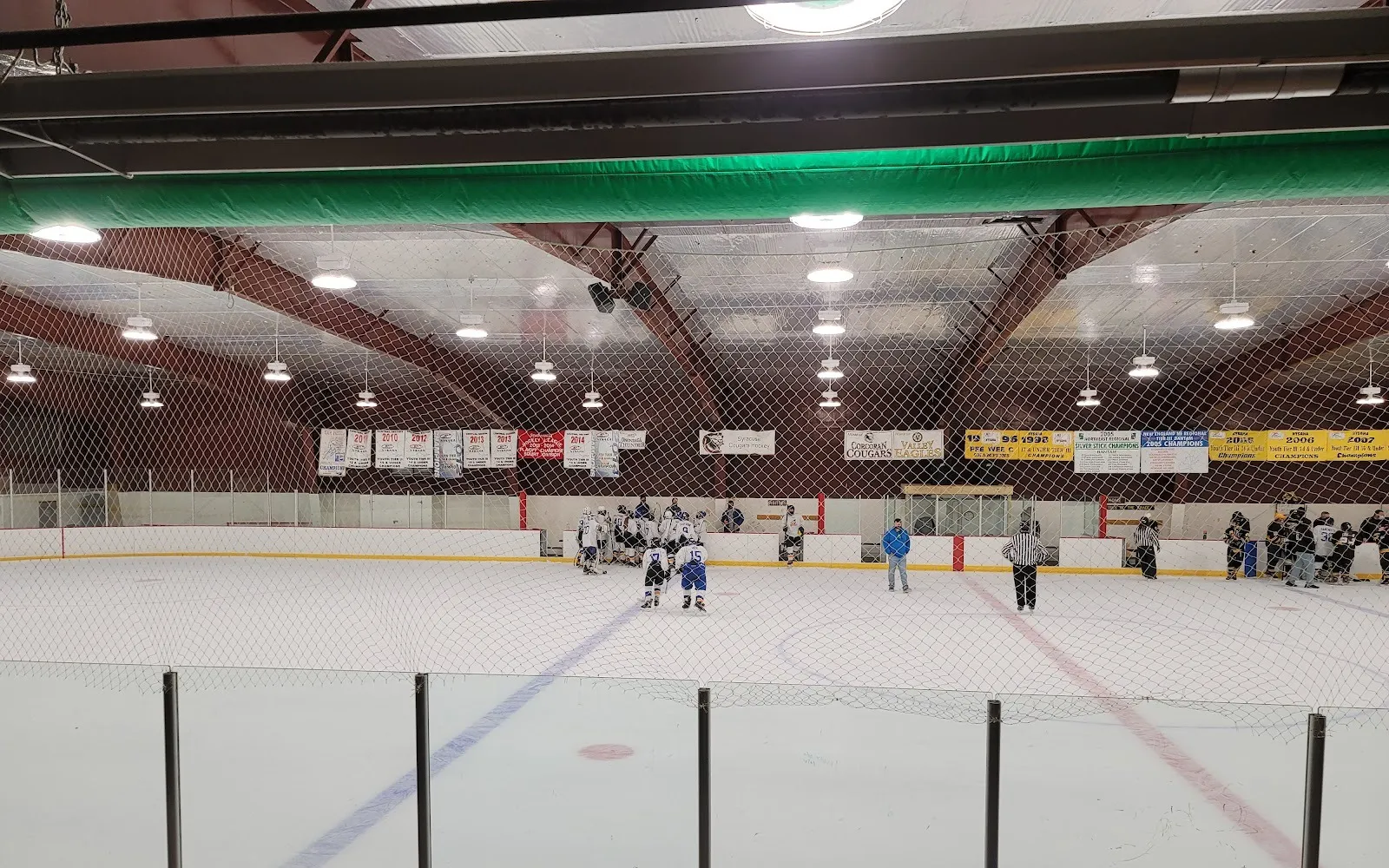 The full sheet seen through the end netting under the dark wooden roof arches, with team banners along the far wall during a youth game. — John Dunn Ice Rink at Meachem Park