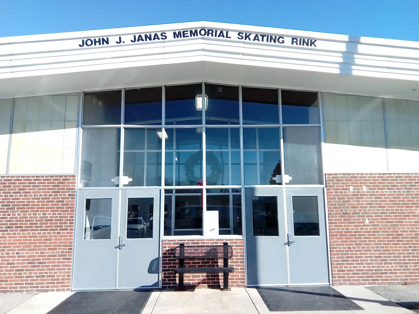 Front entrance of the brick building with a white marquee reading "JOHN J. JANAS MEMORIAL SKATING RINK" above glass double doors and a bench out front. — John J. Janas Memorial Rink