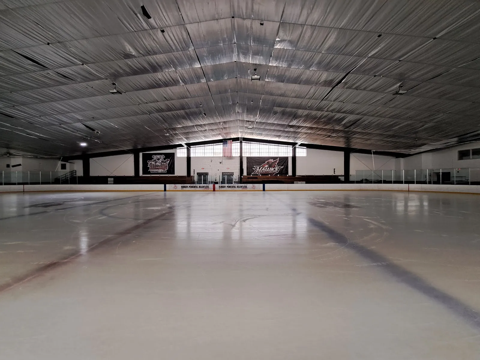 Empty ice sheet viewed rink-side toward the far boards, with "Bulldogs Hockey Club" and "Hawks" team banners on the end wall and a metal-roofed interior; no people on the ice. — John J. Janas Memorial Rink