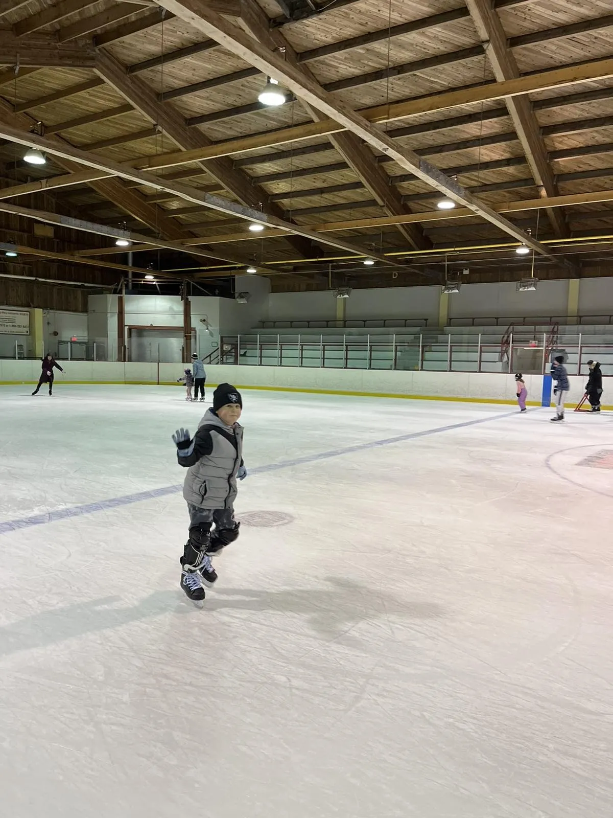 A skater on the ice in a portrait view, a timber-ceilinged hall with players far across the sheet behind. — John Levi Community Centre (Almonte)