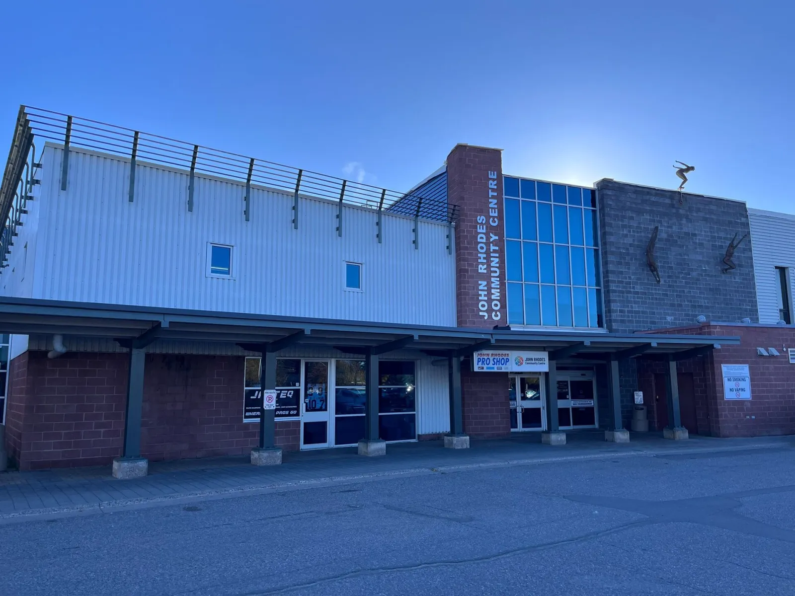 El John Rhodes Community Centre, en Sault Ste. Marie: un edificio bajo de ladrillo y revestimiento metálico con una torre de escalera acristalada, su nombre en la fachada y una marquesina a lo largo de la entrada. — John Rhodes Community Centre