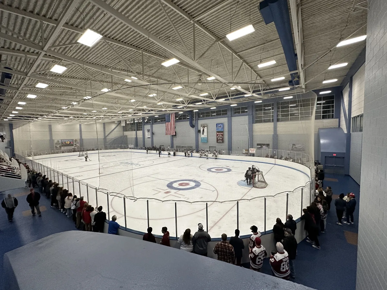 La arena de hielo vista desde el barandal para espectadores, con círculos de curling en la pista y el público a lo largo de las vallas. — Jones Center Ice Arena