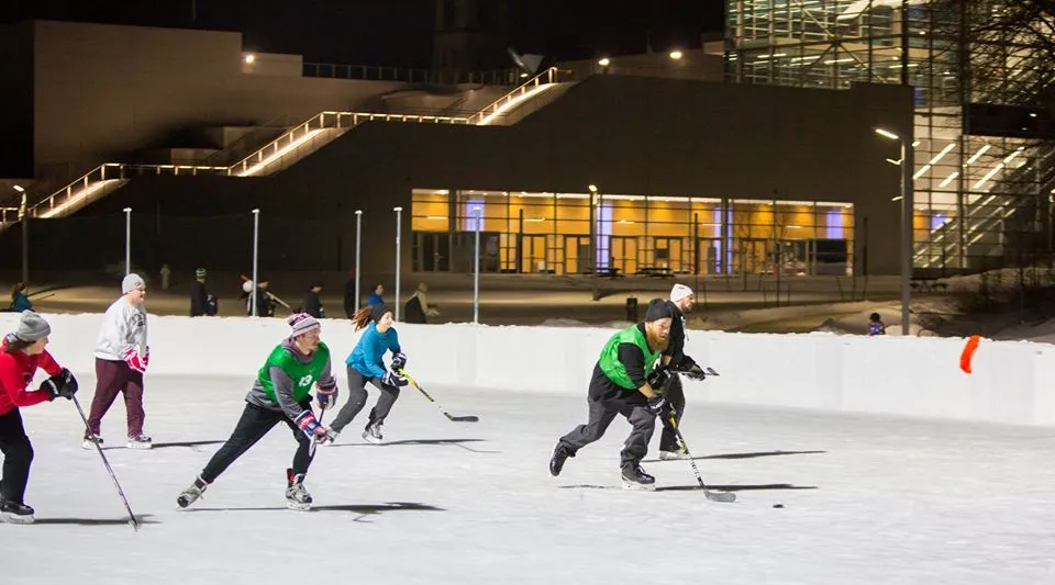 Un partido de hockey en la pista de Jones Park en Appleton, con jugadores repartidos por el hielo y un edificio iluminado detrás de la valla. — Jones Park Hockey and Family Rinks