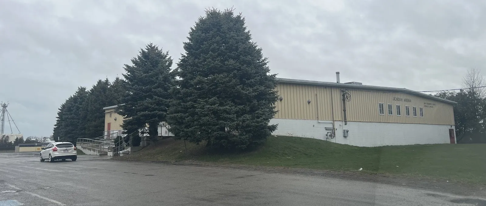 The tan-sided Jordan Lions Arena on Fourth Avenue in Jordan, partly screened by tall spruce trees on an overcast day — Jordan Lions Arena
