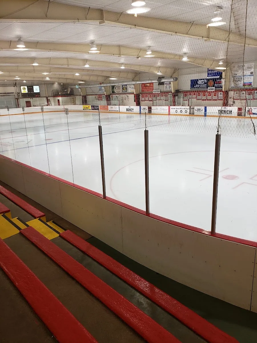 The Jordan arena ice from the red-and-yellow wooden bleachers, with the boards and banners along the far side. — Jordan Lions Arena