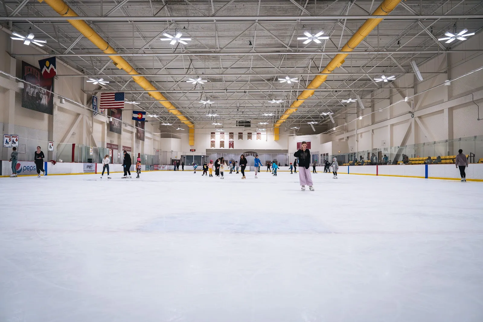 The full sheet from a corner at ice level, with yellow-painted roof trusses, flags and Pioneers banners along the far wall, and a few skaters in the distance. — Joy Burns Arena