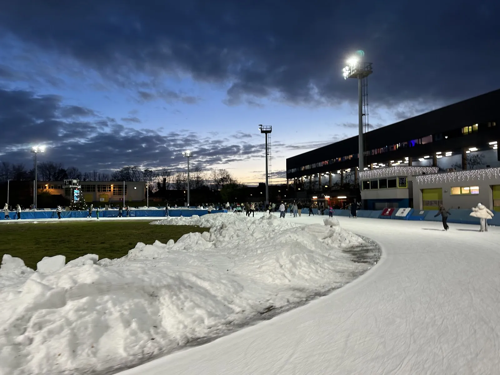 Ein geschwungener Abschnitt der Freiluft-Eisbahn in der Dämmerung, mit einem Schneewall an der Innenseite, Flutlicht und dem dahinter aufragenden Hallengebäude. – JOYNEXT Arena