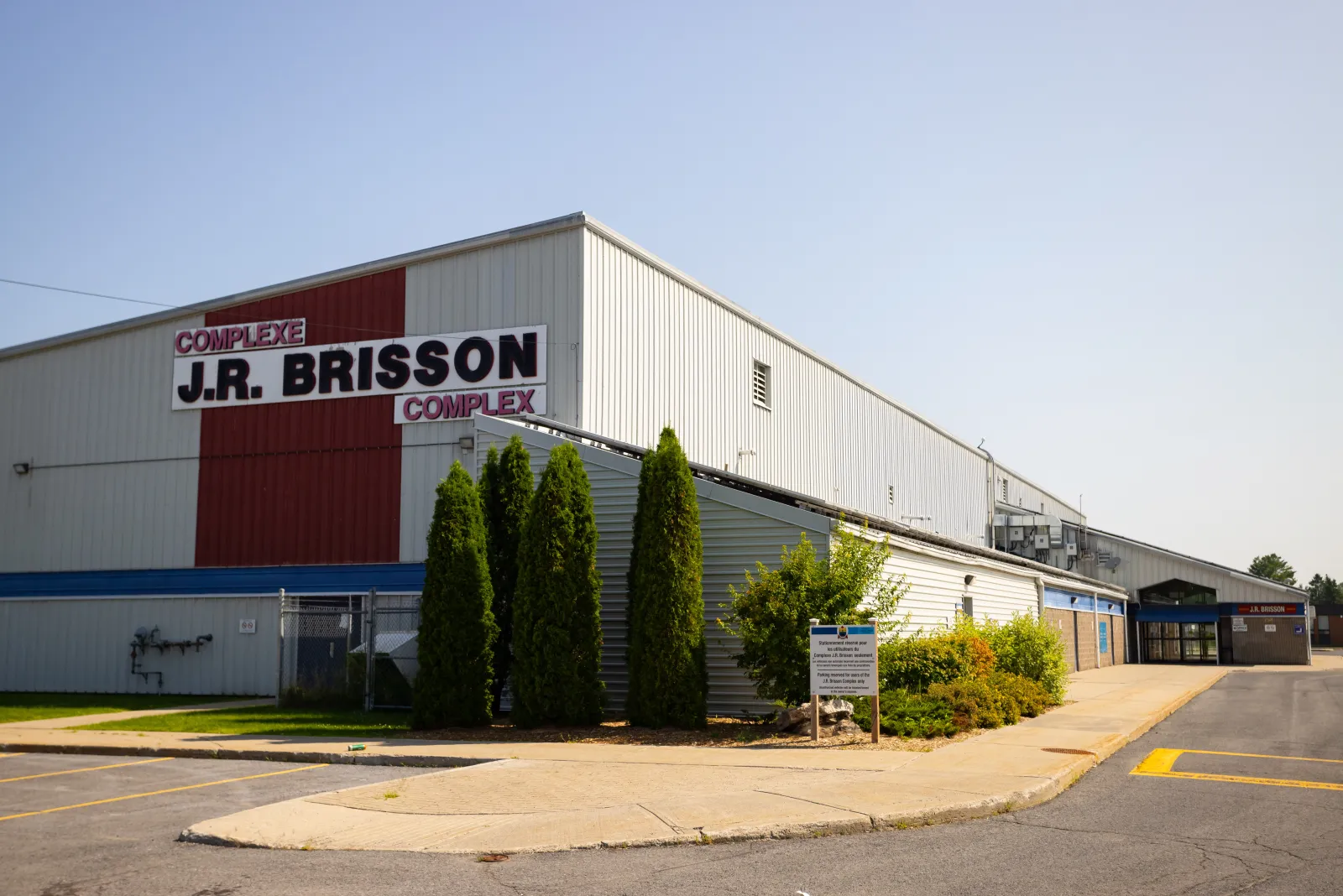 La fachada blanca y roja del J.R. Brisson Complex con su nombre en el muro y una fila de cedros junto a la entrada. — J.R. Brisson Complex (Casselman)