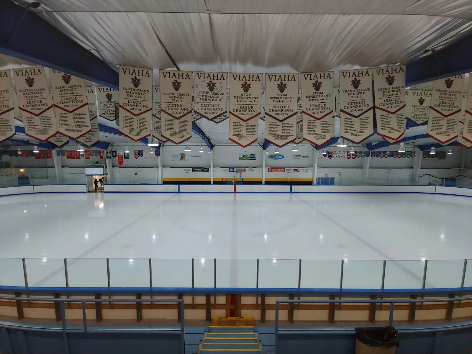 A wide empty sheet seen from behind the glass, a Juan de Fuca banner wall and wooden seating above the far boards under a bright ceiling. — Juan de Fuca Arena
