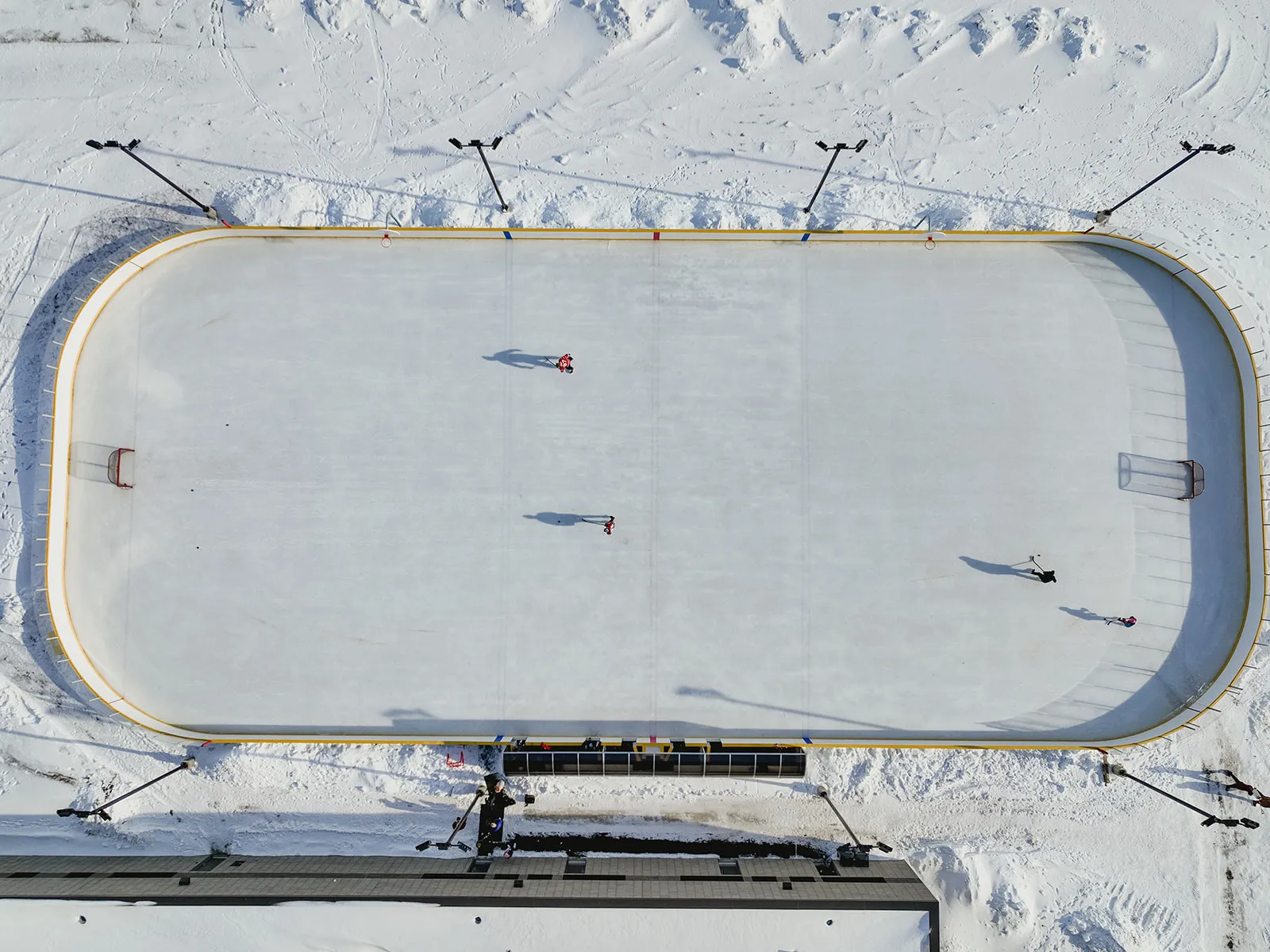 Aerial view of the Jubilee Park rink in Weyburn: a boarded ice sheet with rounded corners in the snow and a couple of skaters on the ice. — Jubilee Park Rink (Weyburn)
