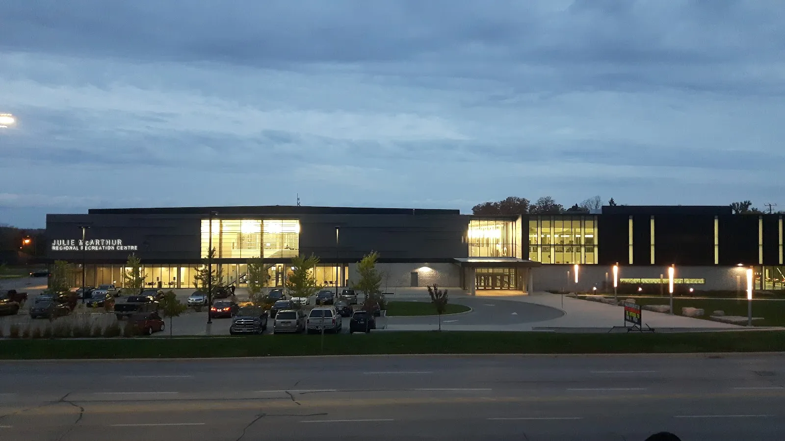 The long, dark-clad Julie McArthur Regional Recreation Centre at dusk with its name on the front wall, lit glass sections glowing and a parking lot in front of it. — Julie McArthur Regional Recreation Centre