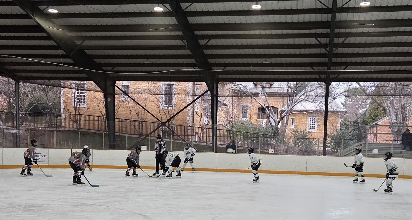 A covered open-sided rink under a dark steel roof with players spread across the ice and a brick building and bare trees beyond the far side. — Jungé Pavilion