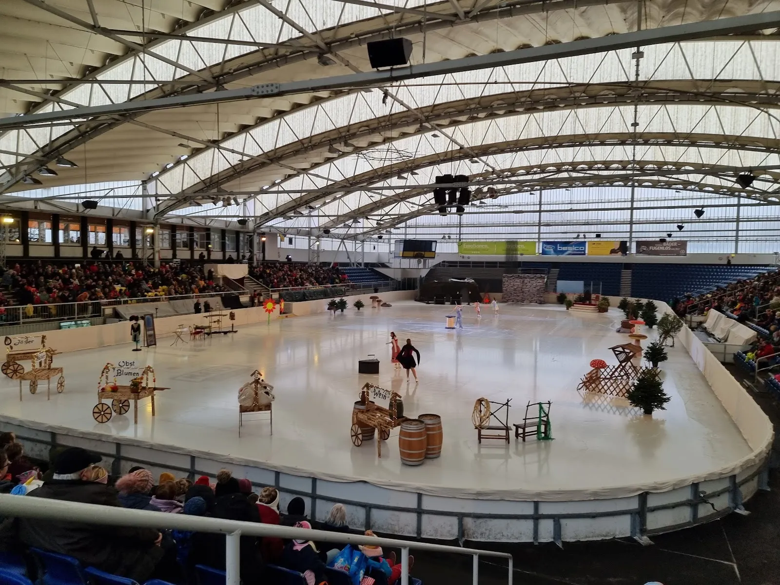 Skaters on the ice of the Jutta Müller Eissportzentrum in Chemnitz, seen from the boards under the arched hall roof. — Jutta Müller Eissportzentrum
