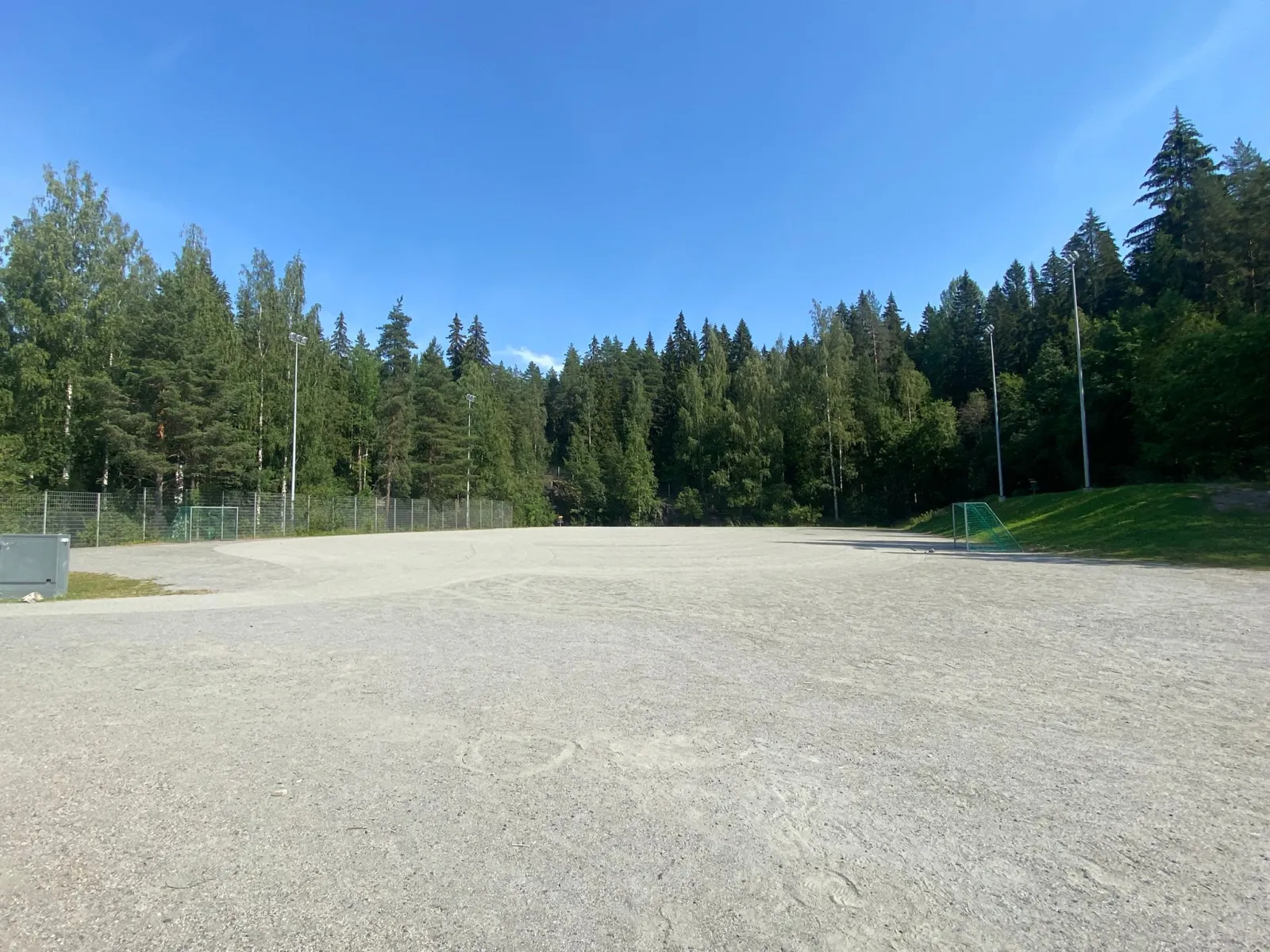 The gravel field at the Jynkkä school in Kuopio, which serves as a skating field in winter, seen in summer with spruce woods behind. — Jynkän koulun luistelukenttä