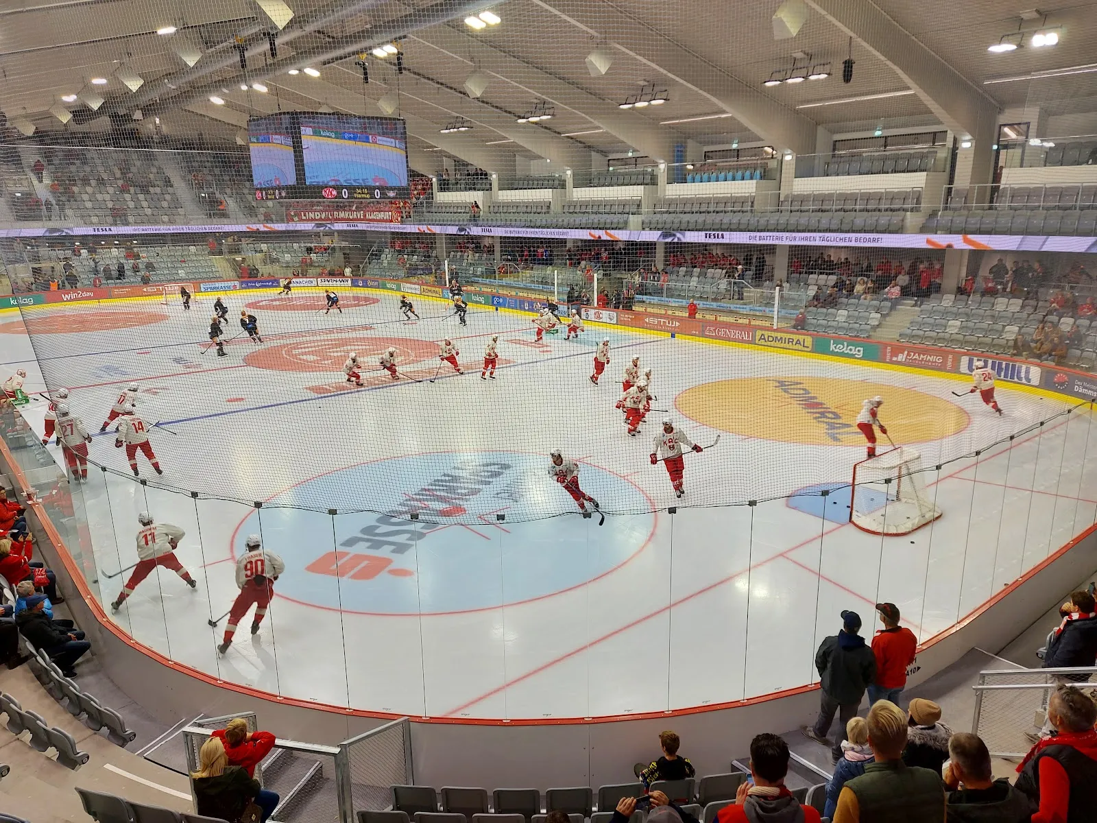 Die Eisfläche der Heidi Horten-Arena im Kärntner Eissportzentrum von den Rängen gesehen, mit weißem Eis, farbigen Kreisen und der Glasbande im Vordergrund, die Spieler klein und über die Fläche verteilt – Kärntner Eissportzentrum Klagenfurt
