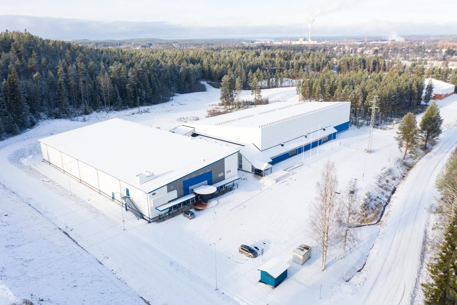 An aerial view of Kajaanin jäähallit in Vimpelinlaakso in winter: the two ice halls stand side by side among snow-covered forest, with the town in the distance. – Kajaanin jäähalli