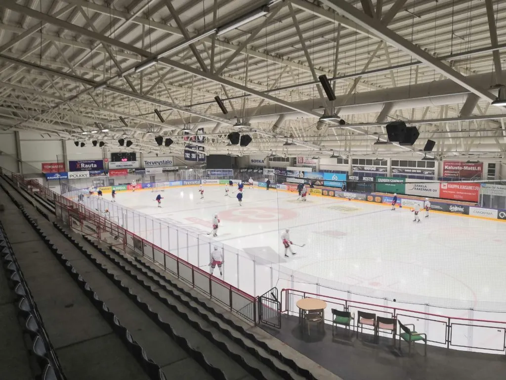 The rink of the competition hall seen from the stands, with skaters spread across the ice, boards covered in advertising and steel roof trusses overhead. – Kajaanin jäähalli