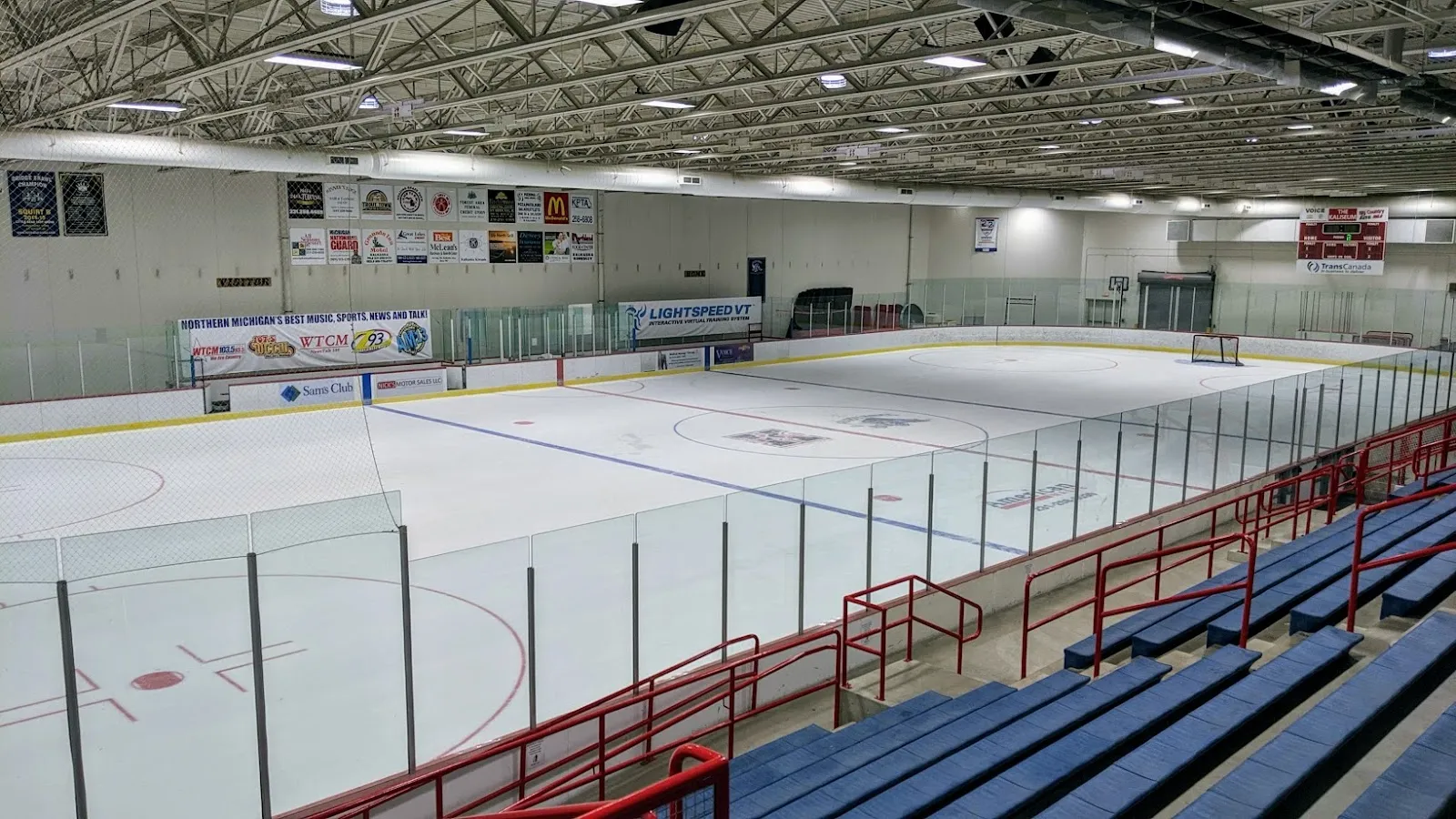 The empty Kaliseum ice sheet under a lit ceiling with boards and stand seating along the side. — Kaliseum Recreation Complex