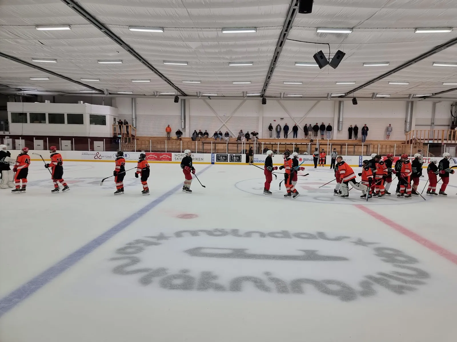 The ice sheet in the hall with a painted blue line and a logo in the ice, a line of red-shirted players at the far boards, and a pale roof with ceiling lights overhead. — Kållereds ishall
