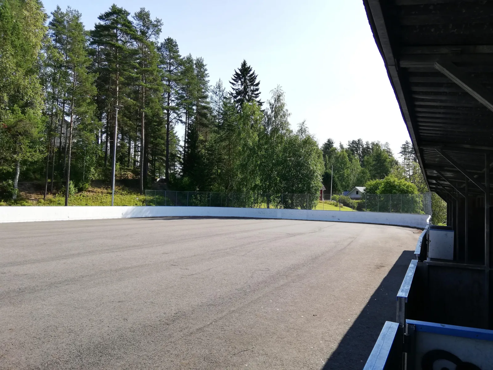 The boarded Kankainen rink in summer, with its dark asphalt surface, boards and a covered players' bench area beside it. — Kankaisen jääkiekkokaukalo