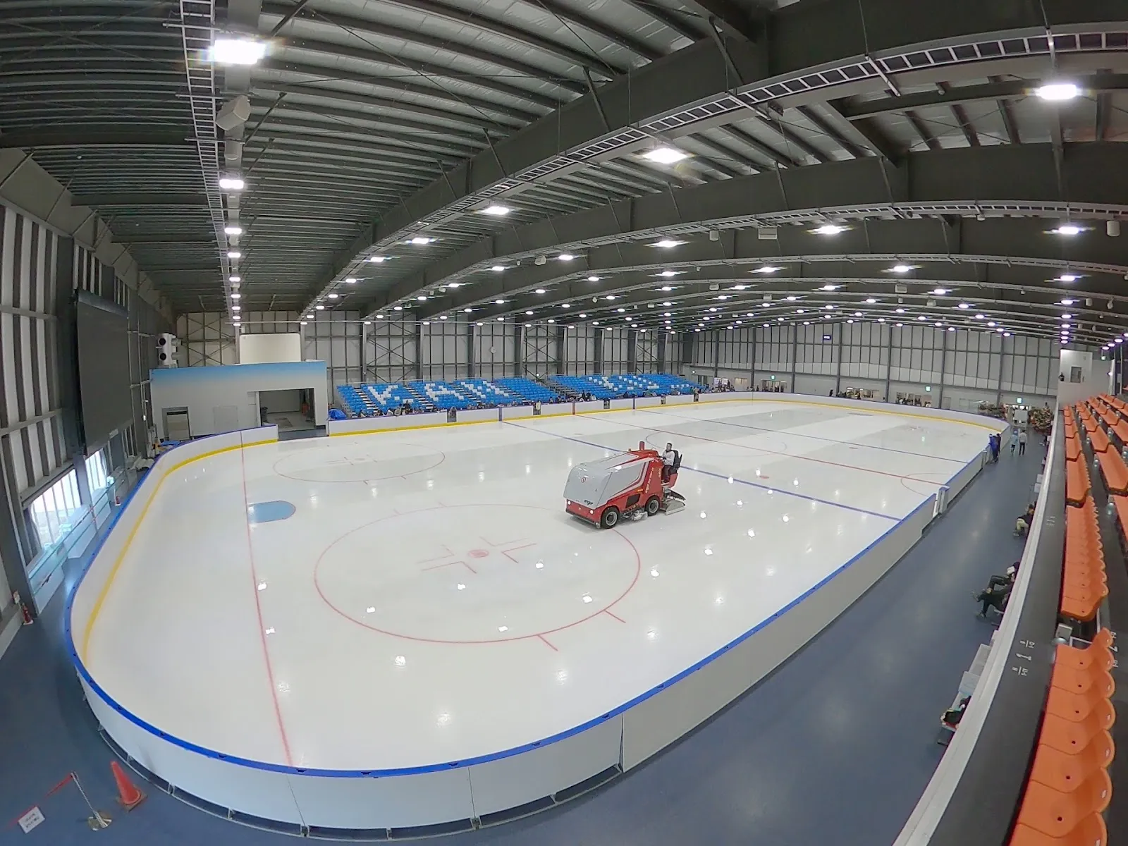 The empty ice sheet of Kanku Ice Arena with a resurfacing machine at centre ice, blue boards and orange seating along the side, seen from the stands — Kanku Ice Arena