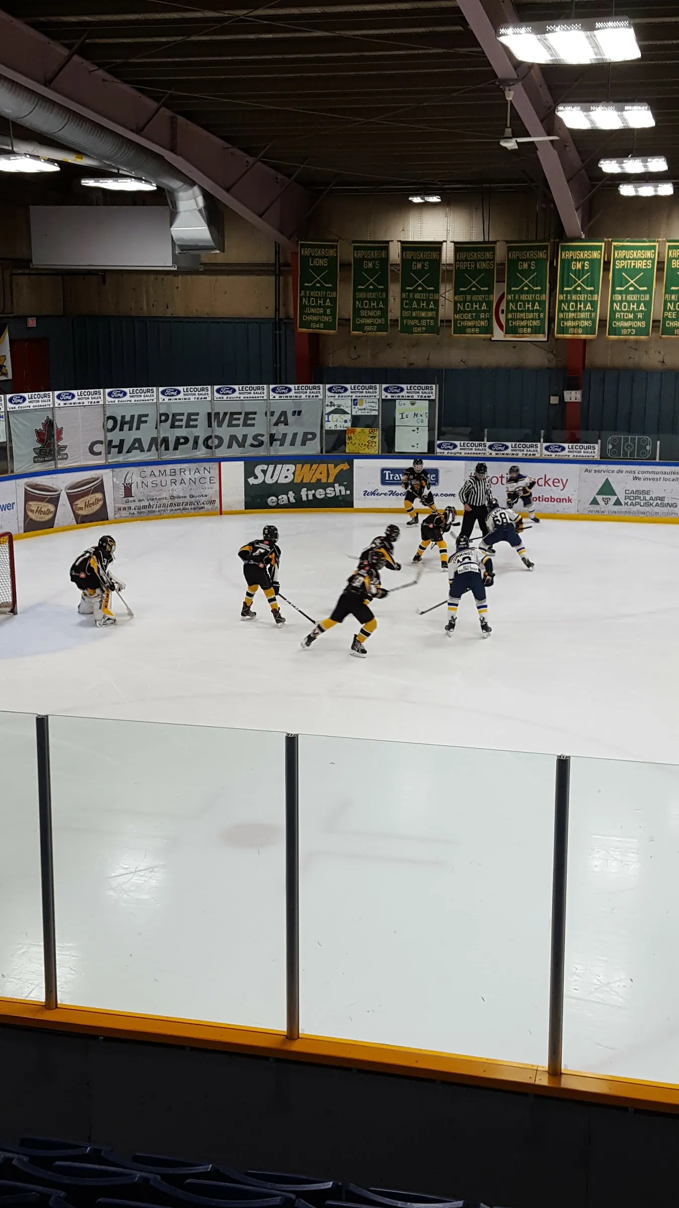 A minor hockey game on the ice at the Kapuskasing Sports Palace, with green championship banners hanging above the boards and sponsor boards along the side. — Kapuskasing Sports Palace