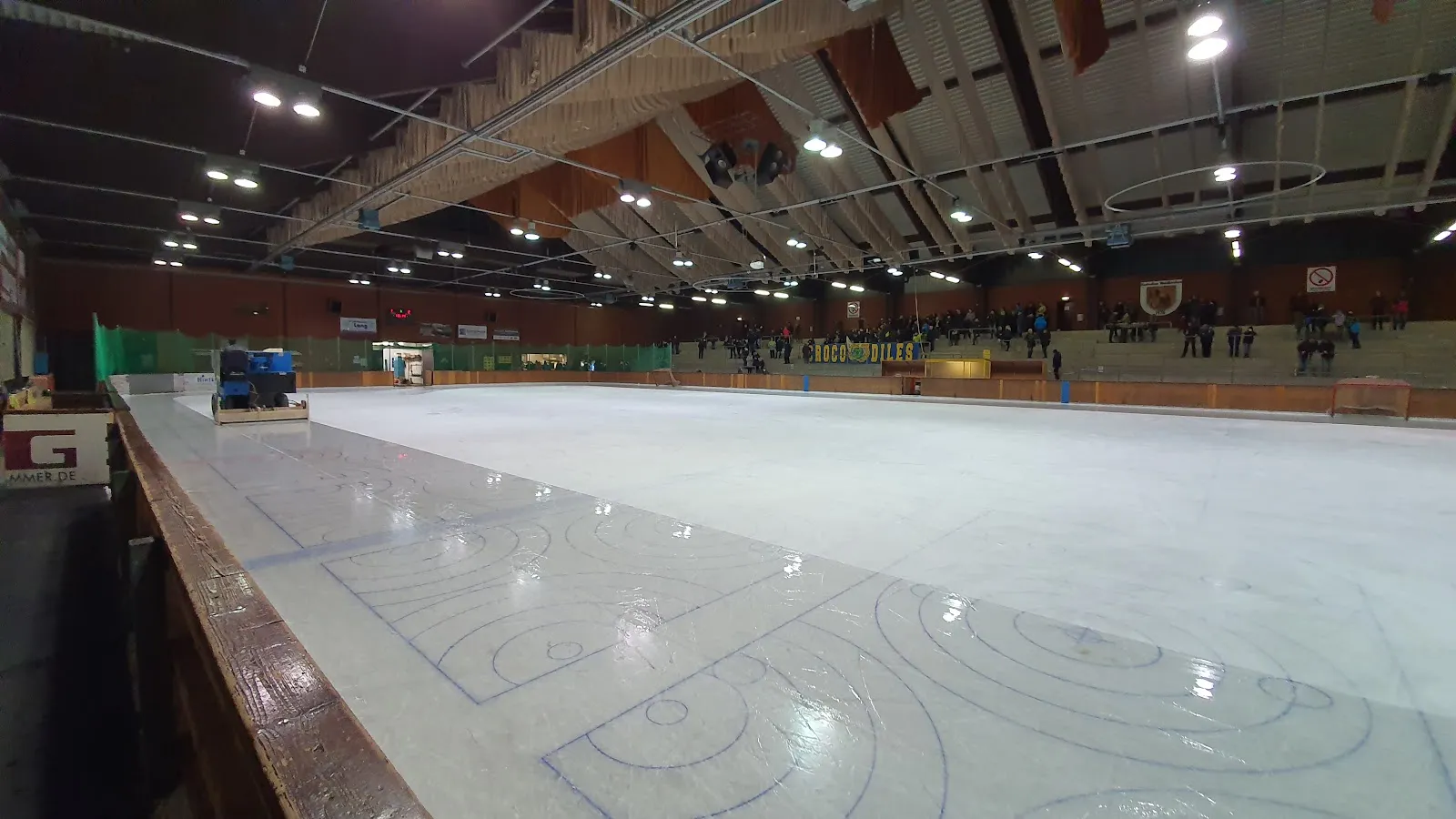 The empty ice surface of the Karoli Eishalle seen along the boards, with the ice resurfacer at the far end and the timber-beamed roof overhead. — Karoli Eishalle