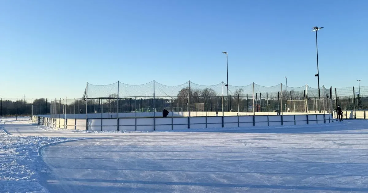 Kartanonkosken liikuntapuiston tekojääkenttä Vantaalla matalassa talviauringossa: laaja jääpinta laitojen ja pallonkeräysverkkojen takana, valomastoja ja yksi luistelija kauempana. – Kartanonkosken liikuntapuiston tekojääkenttä (Vantaa)