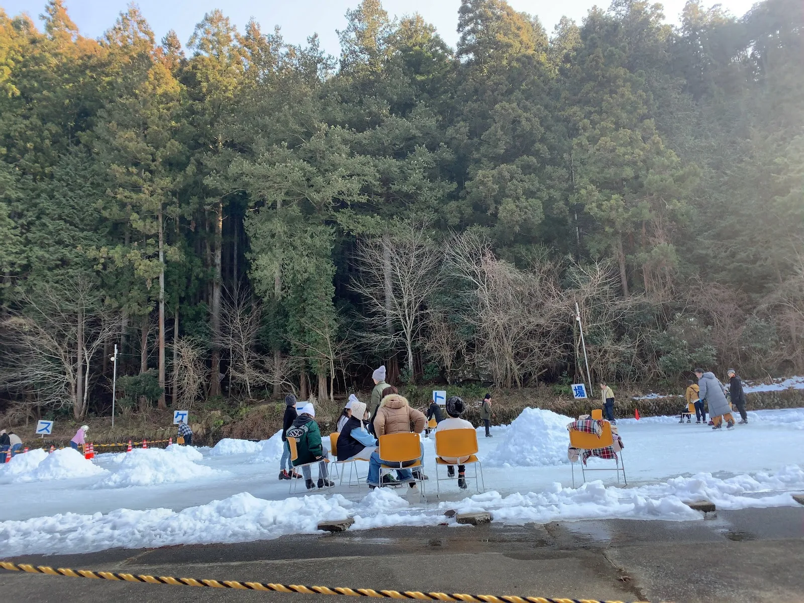 Skaters resting on chairs at the snowy edge of the natural-ice rink, with banked snow, small signs and a dense wall of cedar and bare trees behind. — Kasa Skate Rink