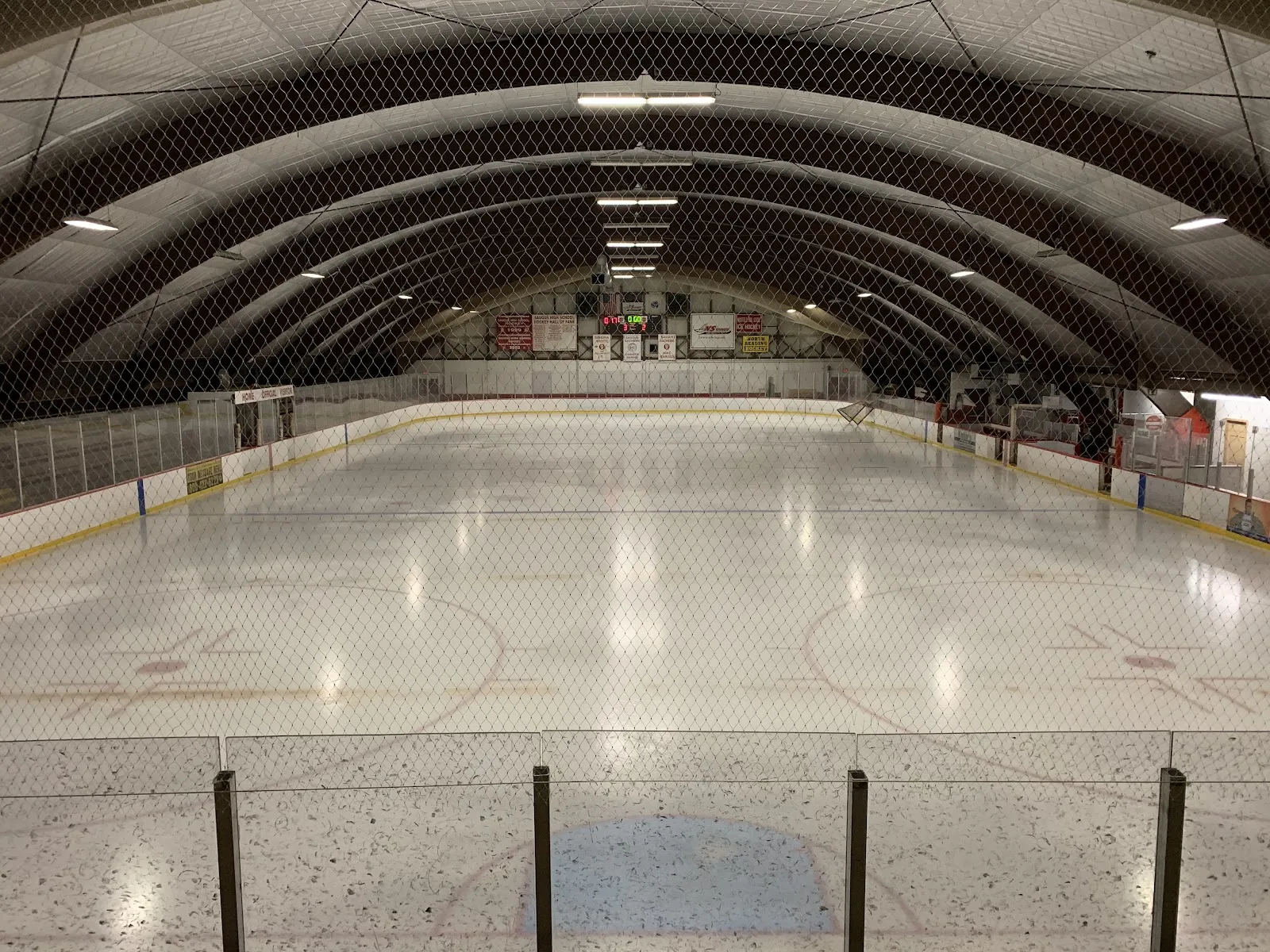 Empty ice sheet under a barrel-vaulted, netted ceiling, with a scoreboard, US and Canadian flags, and Saugus High School Hockey Hall of Fame championship banners on the end wall. — Kasabuski Memorial Rink
