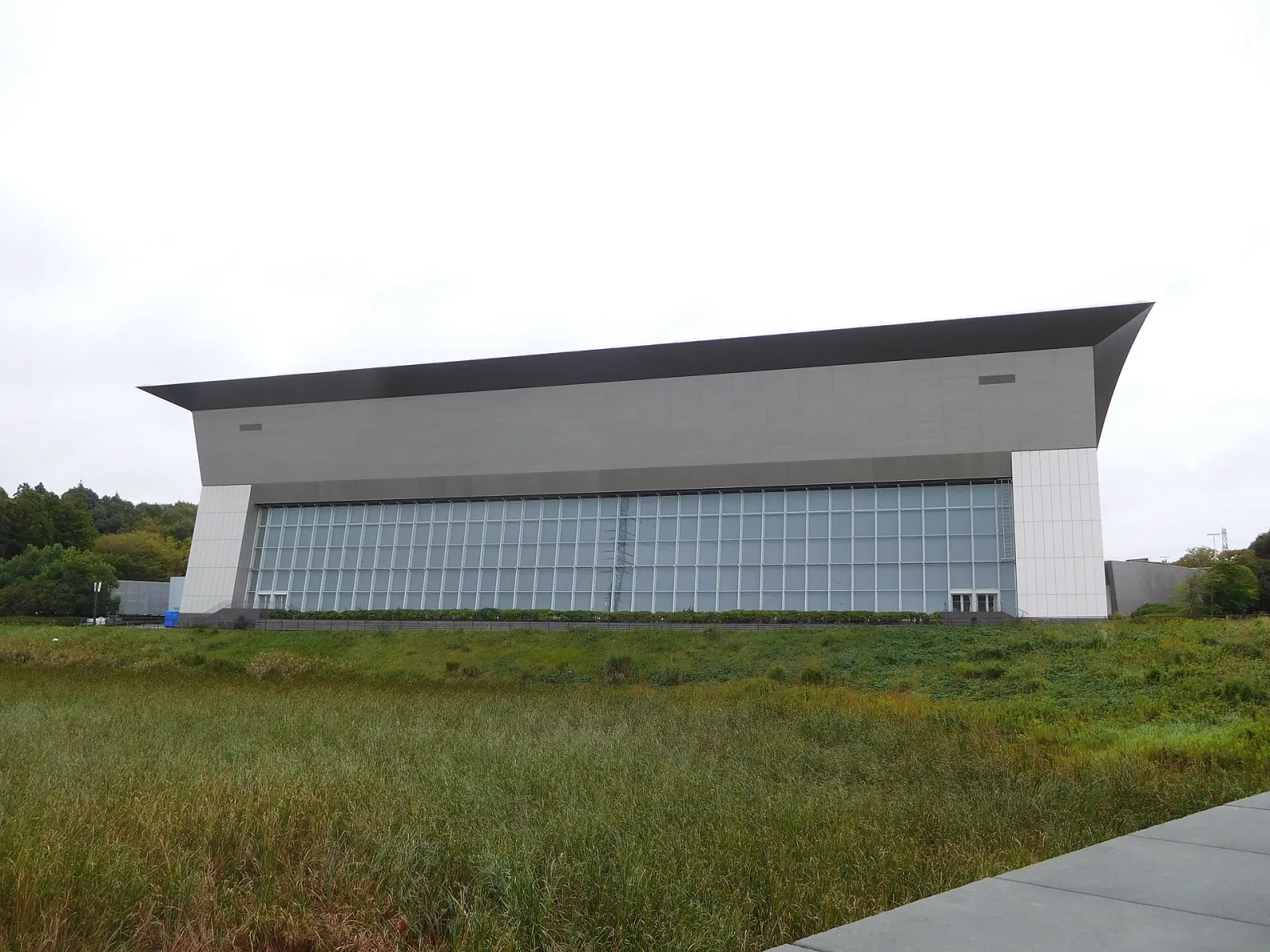 The long glass-fronted hall of the Yamashin Swimming Arena at Kasamatsu Sports Park under an overcast sky, with grass in the foreground. — Kasamatsu Sports Park Ice Arena (Yamashin Swimming Arena)