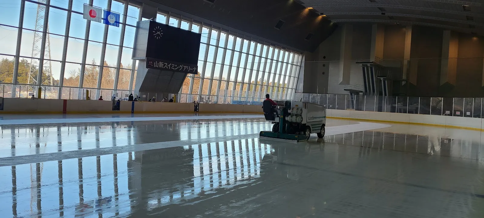 The ice at the Yamashin Swimming Arena with the resurfacer at work in front of a tall glass wall and the scoreboard. — Kasamatsu Sports Park Ice Arena (Yamashin Swimming Arena)