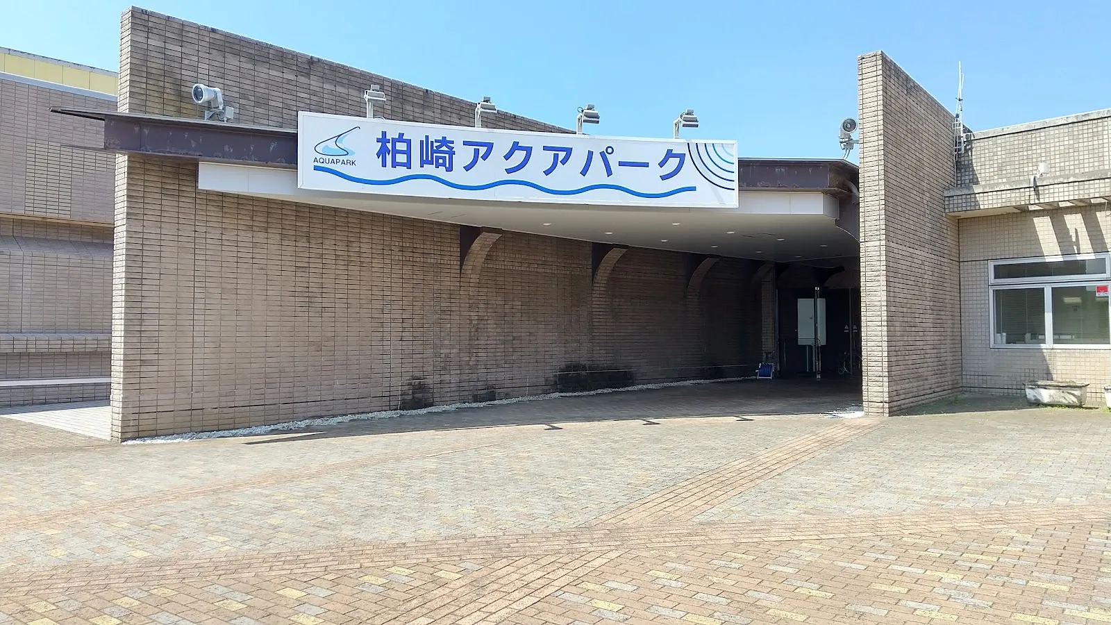 The brick-faced entrance of the Kashiwazaki Aqua Park with its blue wave-logo sign over a paved forecourt under a clear sky. — Kashiwazaki Aqua Park Ice Rink