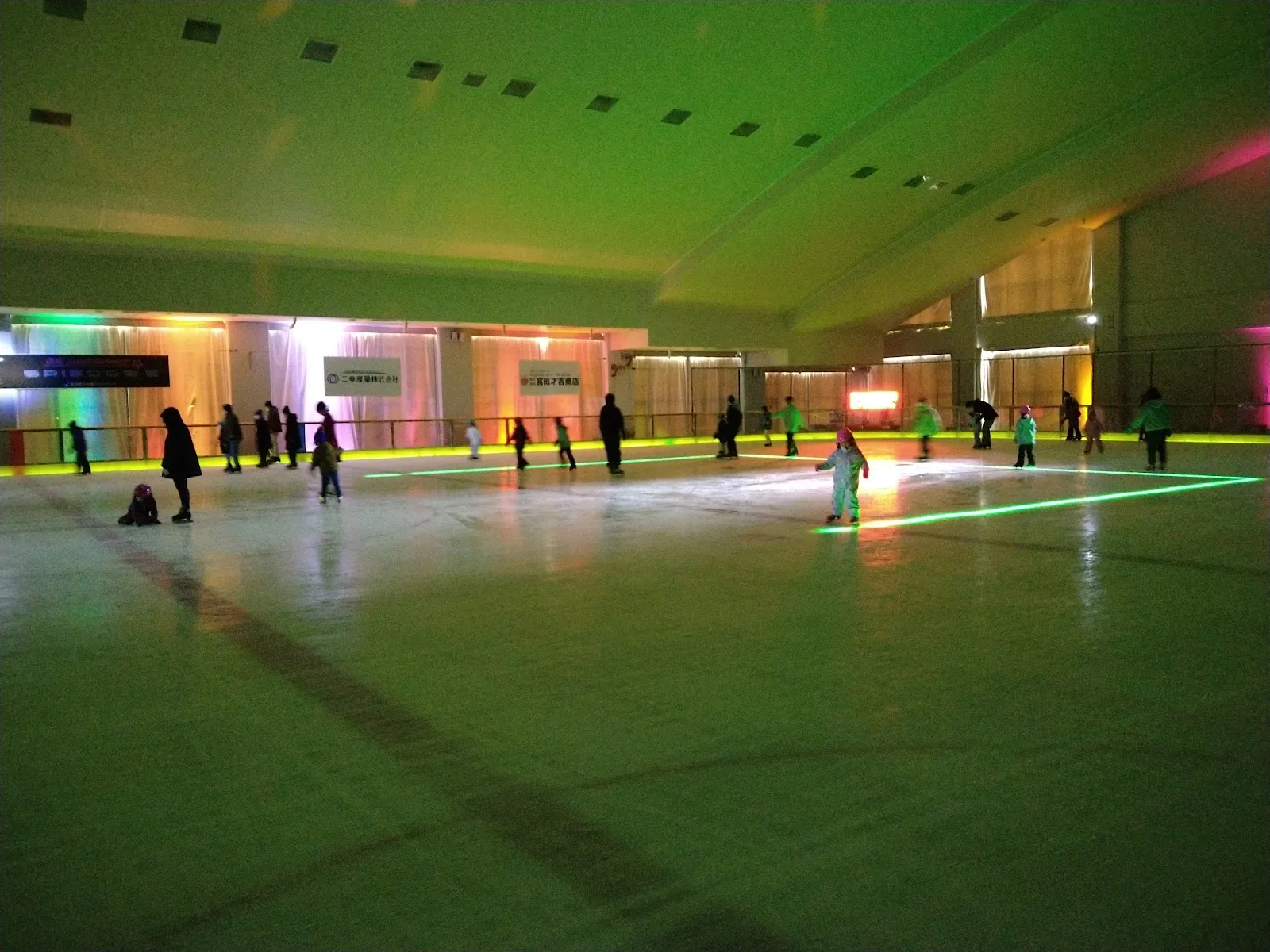The winter ice rink in the Kashiwazaki Aqua Park hall during an illuminated session, with skaters spread across the ice and coloured lights along the far wall. — Kashiwazaki Aqua Park Ice Rink