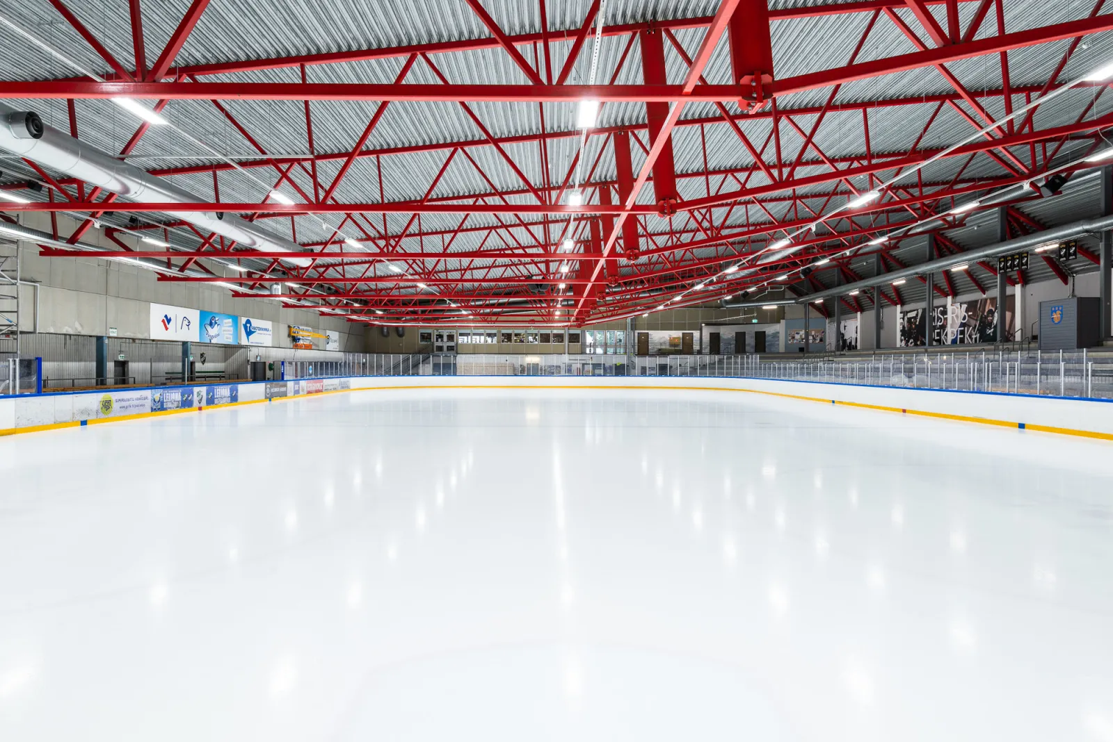 The empty ice sheet of Kauniaisten jäähalli seen from the end of the rink, under a roof of red steel trusses with the stands and boards along the far side. – Kauniaisten jäähalli