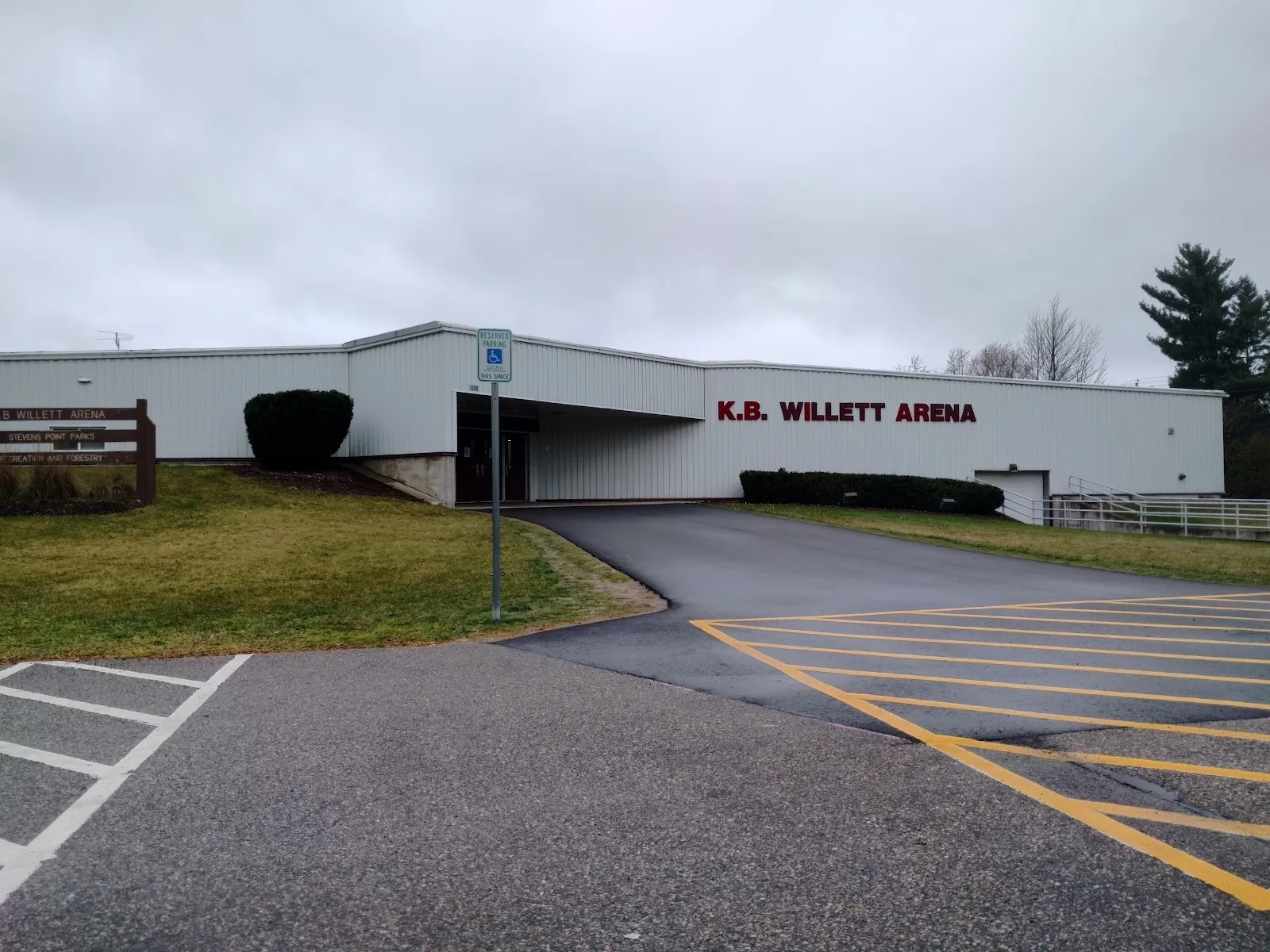 Front of K.B. Willett Arena on an overcast day, white metal-panel building with red K.B. WILLETT ARENA lettering, a covered entrance breezeway, handicap parking sign, and a Stevens Point Parks Recreation and Forestry entrance sign on the lawn. — K.B. Willett Ice Arena