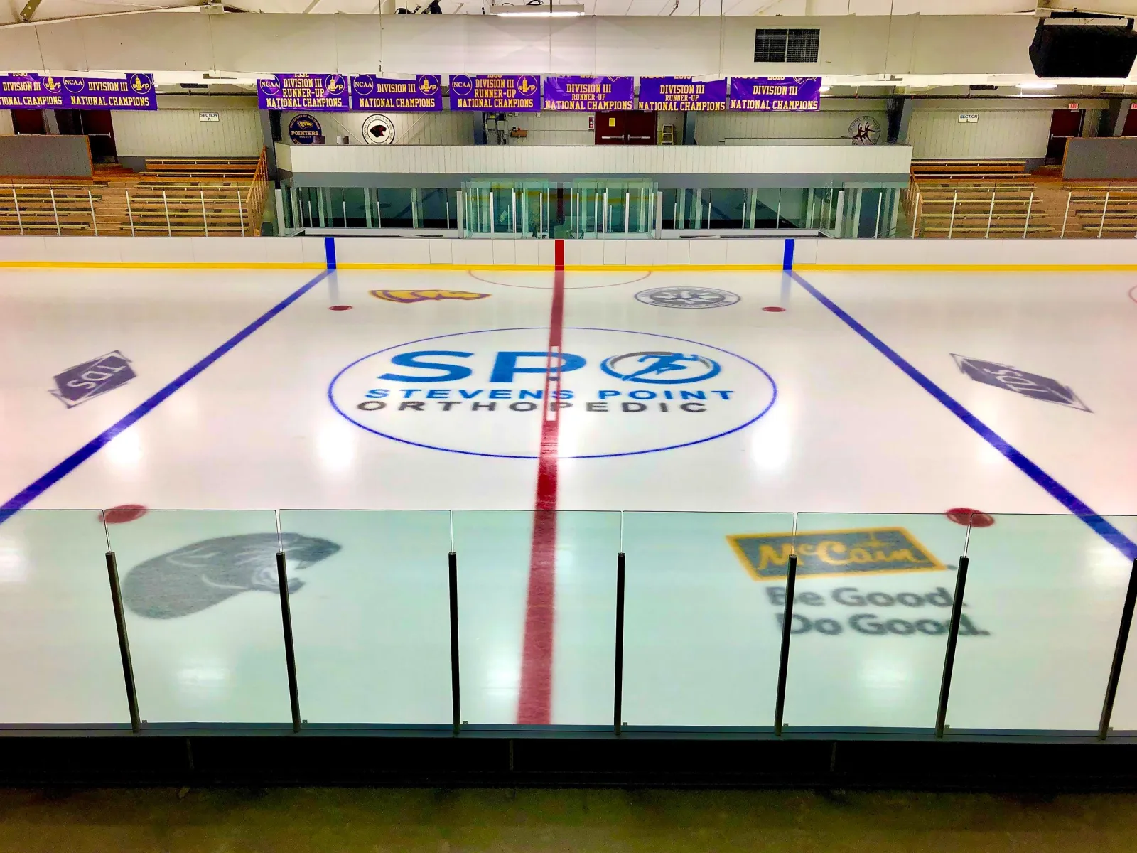Empty ice sheet viewed from an upper corner, with blue and red lines, center-ice sponsor logo, championship banners hanging from the ceiling, and wood bleacher seating along the boards. — K.B. Willett Ice Arena