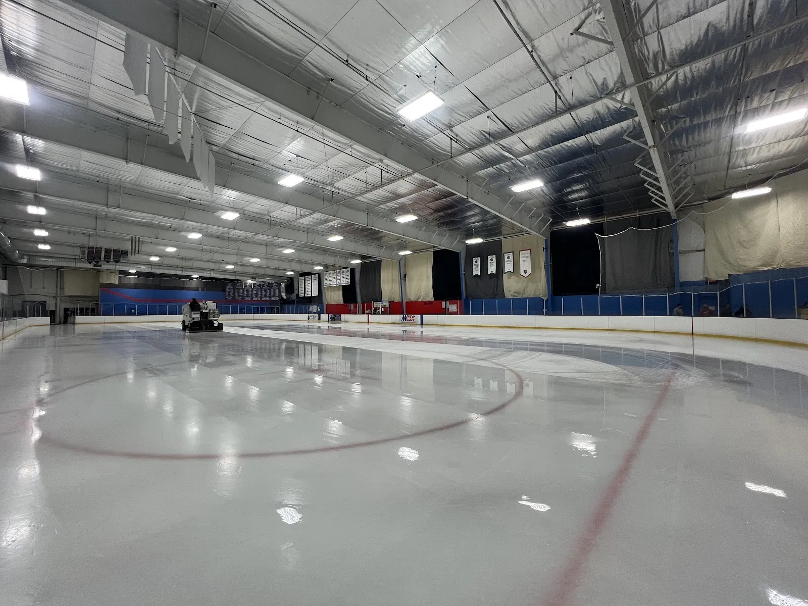 An empty sheet seen from the end, a faceoff circle and red line on white ice under a steel-trussed roof with dark blue boards along the far side. — KC Ice Center