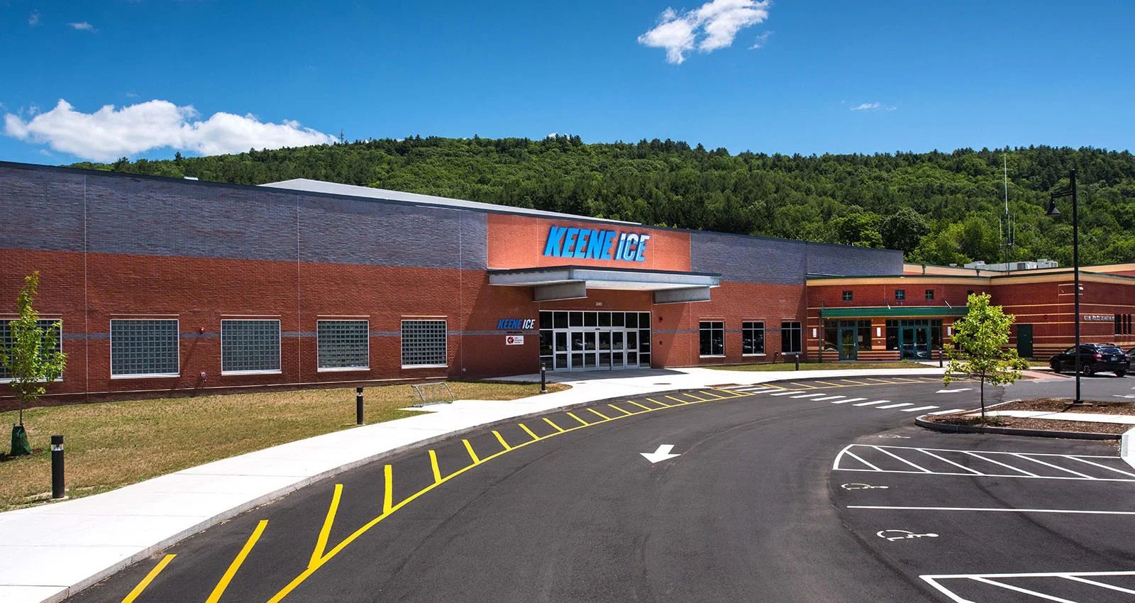 The red-brick Keene ICE building and entrance beside a curving drive, with wooded hills behind. — Keene Ice
