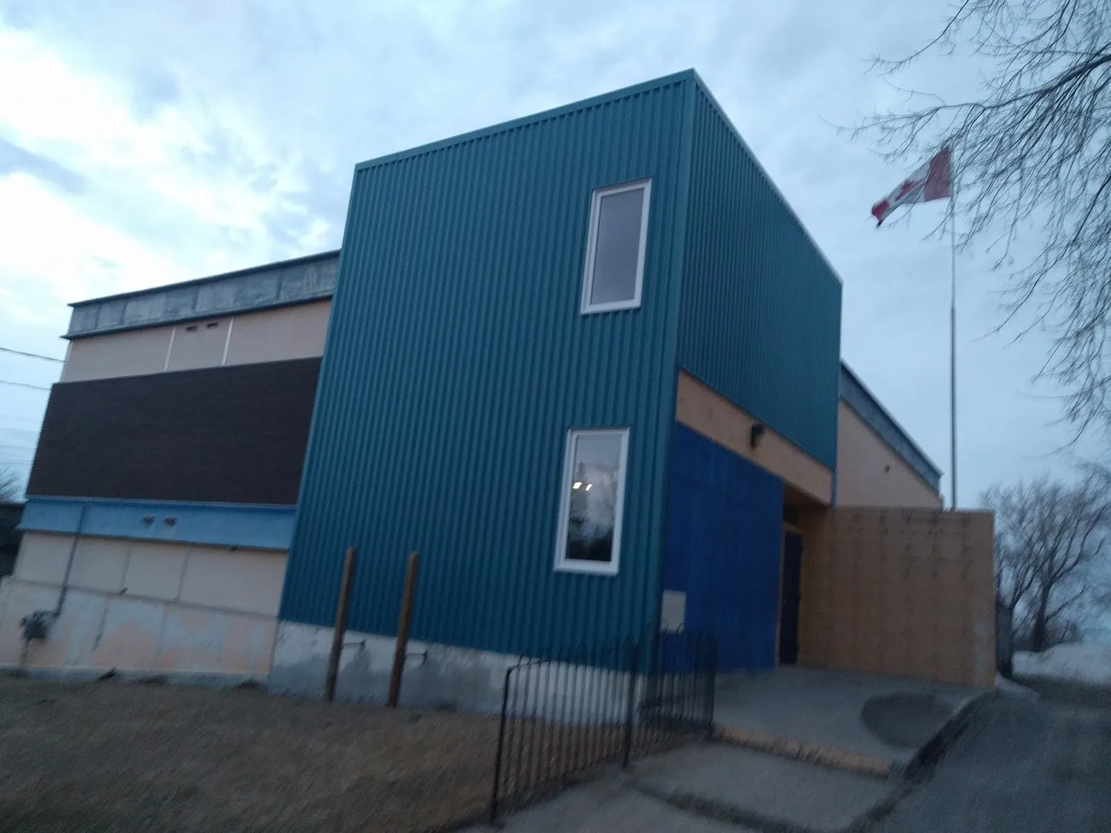 The blue-green corrugated entrance tower of the Keewatin arena building with a tall flagpole flying the Canadian flag, a fence and bare trees at dusk. — Bowman Electric Keewatin Memorial Arena