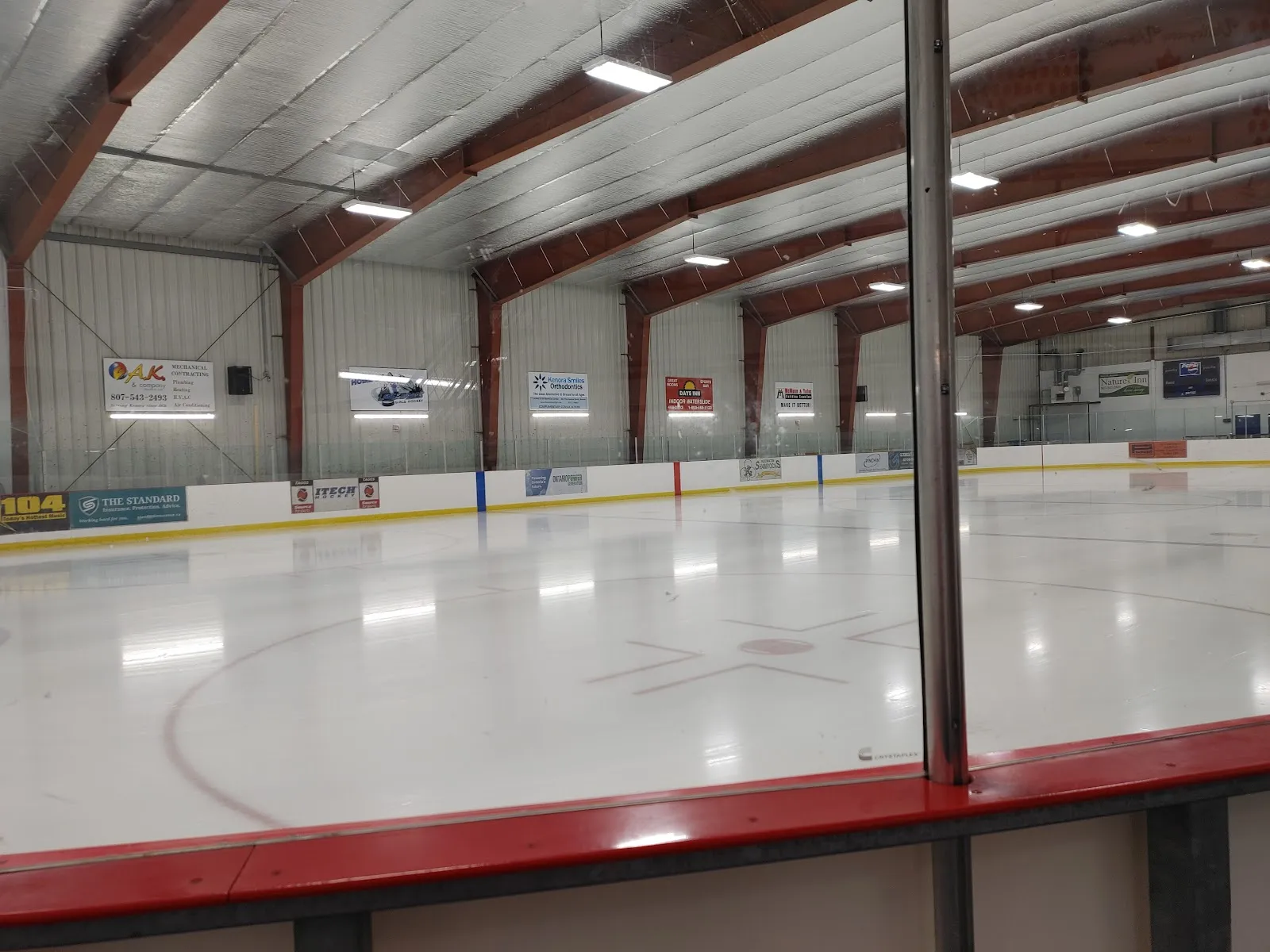 El hielo vacío desde las vallas, bajo las vigas rojas del techo y los letreros de patrocinadores. — Bowman Electric Keewatin Memorial Arena