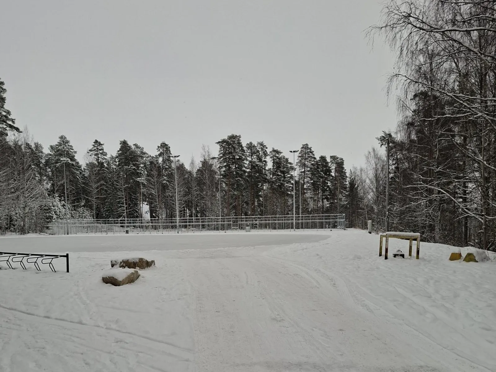 A snow-covered skating field at Kelloniemi seen from a rise, with lamp posts, a bike rack and forest around it. — Kelloniemen luistelukenttä